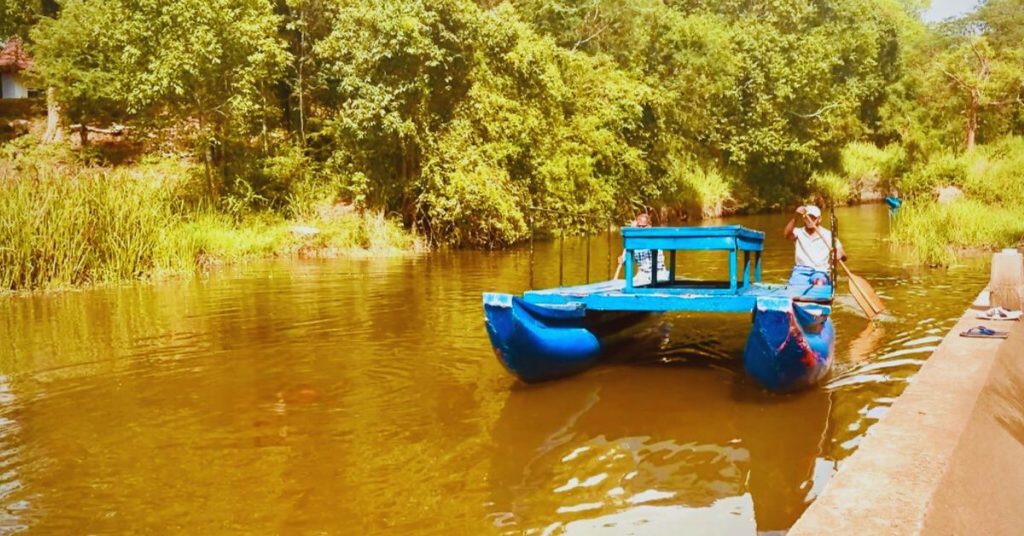 Traditional catamaran gliding across Hiriwadunna Lake in Sri Lanka, surrounded by lush greenery and peaceful village scenery.